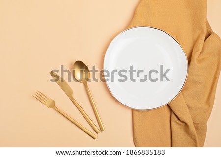 Empty white plate, golden cuterly and cotton napkin on beige backdrop. Food background for menu, recipe book. Table setting. Flatlay, top view, mockup
