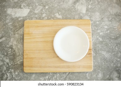 Empty White Plate On The Right Side Of A Wooden Cutting Board On Top Of White And Gray Granite Counter Top. Vertical, Overhead Image With Copy Or Text Space. 