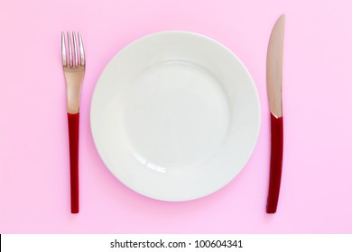 Empty White Plate With Red Handled Fork And Knife On Pink Background. Clean And Simple With Copy Space. Top View, Looking Down.