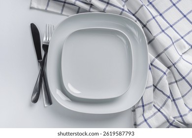 Empty white plates with cutlery and kitchen towel. Minimalist top view of clean white plates, fork, knife, and checkered kitchen towel on a light background, ready for serving.