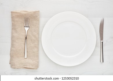 Empty White Round Ceramic Plate, Fork, Knife And Napkin On White Wooden Table. Top Down View. 