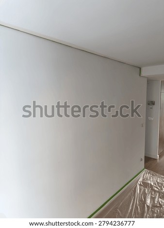 Empty white wall corner prepared for painting with green painters tape along ceiling line and baseboard plus plastic floor cover. Home renovation preparation and DIY repair concept.