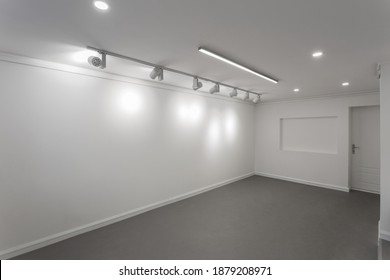 Empty White Wall Without Frames In Gallery Room. Illuminated Space For Art Exposition. Gallery Lighting, Spotlights No People. Showroom With A Dark Gray Self-leveling Floor. 