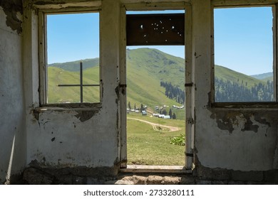 Empty window frames reveal a rural Batumi mountain settlement, tin roofs, winding dirt road, ridge with dark conifers, clear summer sky, bullet marks, metal cross bar.