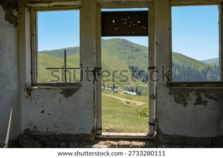 Empty window frames reveal a rural Batumi mountain settlement, tin roofs, winding dirt road, ridge with dark conifers, clear summer sky, bullet marks, metal cross bar.
