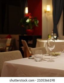 Empty Wineglass And Drinking Glass With Stainless Steel Tabelware On White Tablecloth With Napkin At Fine Dining Restaurant At Night