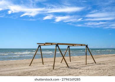 Empty wooden canopy frame setup on a sandy beach next to the sea under a bright blue sky with wispy clouds in summer.
