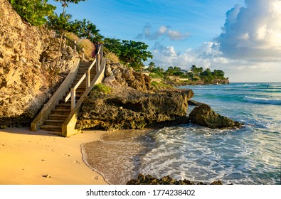 Empty Wooden Staircase Leads Down To An Idyllic White Sand Beach In Barbados. Golden Evening Sunbeams Shine On The Rocky Shore Near A Popular Surf Spot In Barbados. Ocean Waves Wash Shore At Sunrise.