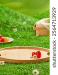 An empty wooden tray for displayed products, decorated with some tomatoes and a daisy, on a green lawn, behind is a tomato dish and a basket. Advertising product photo with a picnic theme.