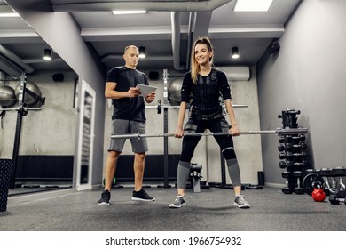 EMS training studio. Female athlete wearing an EMS suit and a trainer in gray and black sportswear lifting a bar while her personal trainer looking at her performance. Electrical muscle stimulation - Powered by Shutterstock