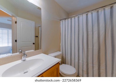 En suite bathroom interior of home with white sink above built in brown cabinet. The clean toilet with closed lid is beside the bathtub and shower covered by white shower curtain.