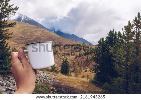 Enamel white mug mockup with forest and mountains valley background. Trekking merchandise and camping geer marketing photo. Stock wildwood photography with white metal cup. 