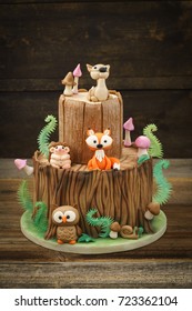 Enchanted forest woodland themed fondant cake with a hedgehog, deer, owl, fox, snail, tree trunk, ferns, mushrooms and leaves on wooden background