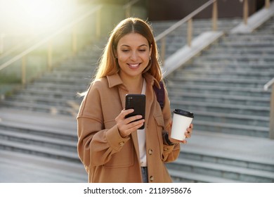 Enchanting blonde young woman waiting for phone message while carrying coffee cup on the street. Stylish girl in spring clothing holding smartphone and take-away cappuccino.