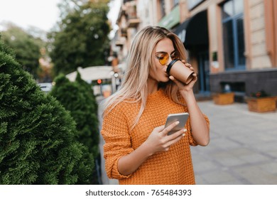 Enchanting blonde young woman waiting for phone message while drinking coffee on the street. Stylish girl in yellow sweater posing on alley holding smartphone and enjoying cappuccino. - Powered by Shutterstock