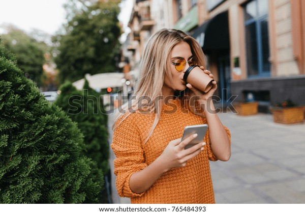 通りでコーヒーを飲みながら 電話を待つ金髪の若い女性を魅惑する スマートフォンを持つ路地でポーズをとり カプチーノを楽しむ黄色いセーターを着たスタイリッシュな女の子 の写真素材 今すぐ編集