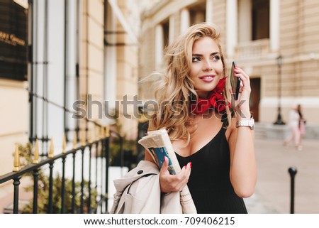 Enchanting tanned female model looking away with inspired smile hanging out outside. Outdoor portrait of laughing blonde woman in stylish black attire holding smartphone in hand.