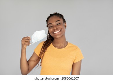 End of coronavirus pandemic. Happy black woman taking off medical mask, feeling free on grey studio background. African American female removing facial covid protection, celebrating end of lockdown