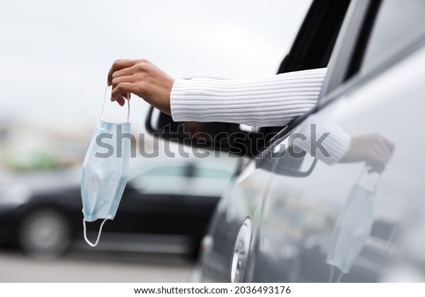 End of covid-19 quarantine, personal transport,\
trip in city by car, new normal and social distance. Young afro\
american woman driver drives car and holds protective mask in her\
hand at open window