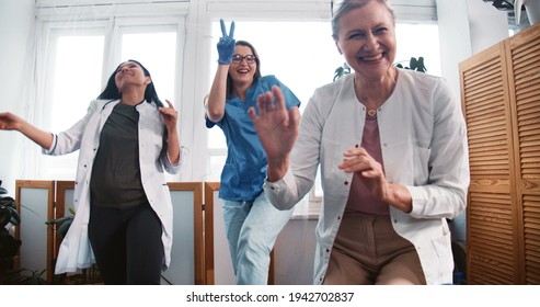End of fighting coronavirus. Zoom out three fun multiethnic happy female doctors dancing together at clinic lab office.