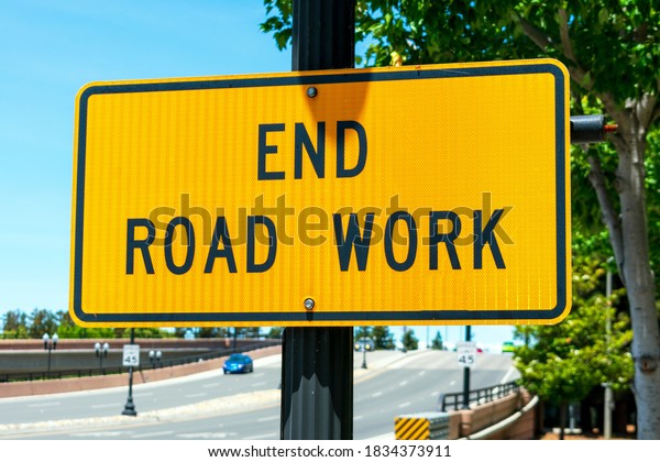 End Road Work reflective\
traffic sign. Background wide urban street going up hill with a few\
cars.