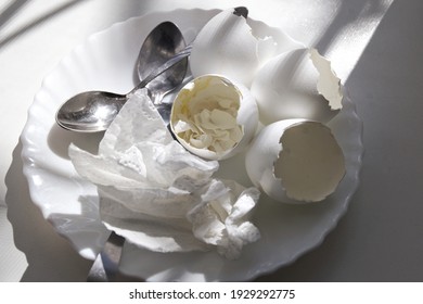 Ending Of Morning Meal: Shells Of Soft-boiled Eggs With The Remains Of The Yolk And The Cooked Egg White, Used Paper Napkin, Teaspoons, All On White Plate
