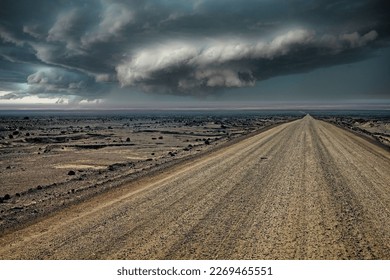 Endless gravel road in monotonous life hostile arid barren surreal gray stone and sand desert landscape, ominous dark rain storm clouds front over horizon 