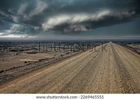 Endless gravel road in monotonous life hostile arid barren surreal gray stone and sand desert landscape, ominous dark rain storm clouds front over horizon 