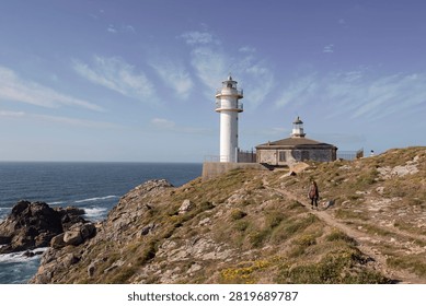Enduring lighthouse overlooking the raw Atlantic cliffs as a solitary hiker advances (Touriñán, Spain: 14-jun-2024)