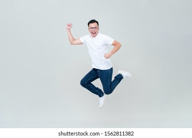 Energetic Asian Man In Casual White T-shirt And Jeans Jumping On Light Gray Background