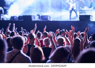 Energetic audience enjoys night concert, raising hands, recording live band on stage with smartphones. Festivalgoers immersed in music, colorful stage lights illuminate excited fans at outdoor event. - Powered by Shutterstock