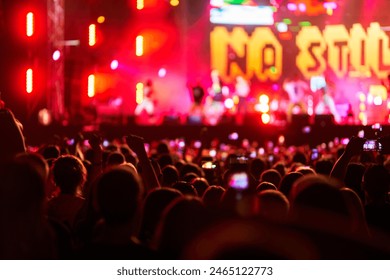 Energetic audience at night music fest, brightly lit stage ahead, concertgoers with hands up enjoy live performance, blurred view creates atmosphere, vibrant festival vibes with excited fans - Powered by Shutterstock