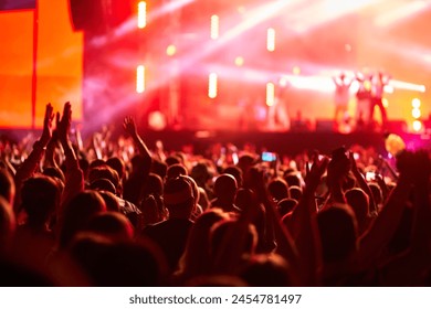 Energetic audience at outdoor live concert at sunset. Fans raise hands, enjoy music at summer festival. Stage lights illuminate excited crowd. Event in full swing with vibrant energy, community vibe.