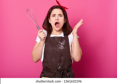 Energetic Emotional Female Wears Dark Brown Messy Apron, White Tshirt, Accessories, Puts Her Hands Up Holding Whisk. Shocked Young Lady Filled With Indignation While Practising Cooking Skills.