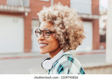 Energetic enthusiastic latin american woman with afro curly crisp hair and eyeglasses listens music via headset, smiling outdoors, looking away. Face shot, natural light.