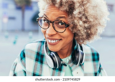 Energetic enthusiastic latin american woman with afro curly crisp hair and eyeglasses listens music via headset, smiling outdoors, looking at camera. Face shot, natural light.