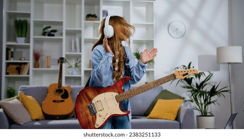 Energetic funny girl with long hair playing electric guitar, sing song and dancing at living room at home. Attractive girl rocker of 11-12 years old playing rock music on electric guitar. - Powered by Shutterstock