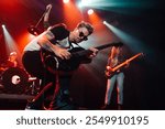 Energetic guitarist concentrating while playing electric guitar during a live concert, illuminated by colorful stage lights, with band members in background