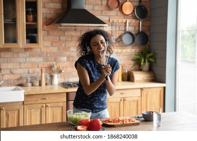 Energetic Overjoyed Young Happy African Ethnicity Woman Wearing Wireless Headphones, Singing Songs In Utensils, Having Fun Entertaining, Enjoying Preparing Food Alone In Kitchen At Leisure Time.