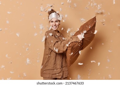 Energetic Pretty Woman Has Pillow Fight In Bedroom Before Going To Bed Wears Slumber Suit And Blindfold Smiles Happily Isolated Over Brown Studio Wall Feathers Floating In Air. Time To Sleep