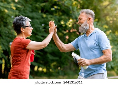 Energetic senior man and woman giving high-five to each other outdoors, happy cheerful elderly people celebrating fitness goals in sunny green park, holding water bottles in hand, side view - Powered by Shutterstock