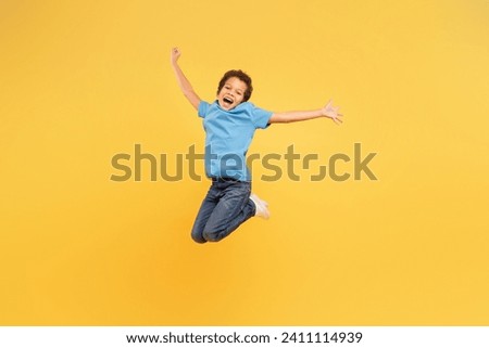 Energetic young boy with bright smile, leaping high with arms spread wide, dressed in blue shirt and jeans against sunny yellow backdrop