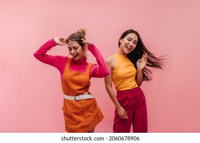 Energetic young two interracial girls dancing side by side and waving hands in studio. Models with light brown and black hair in light spring orange and pink clothes move to music with smile on face. - Shutterstock ID 2060678906