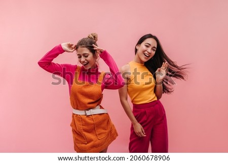 Energetic young two interracial girls dancing side by side and waving hands in studio. Models with light brown and black hair in light spring orange and pink clothes move to music with smile on face.