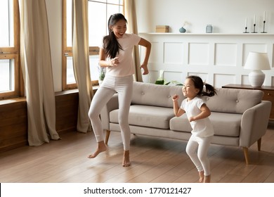 Energetic young vietnamese ethnic mother enjoying playtime with funny crazy asian child daughter, dancing to favorite disco music barefoot in modern living room, hobby weekend pastime indoors.