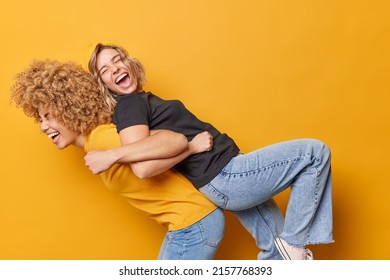 Energetic young women lean at backs of each other give piggy back ride dressed in casual t shirt and jeans feel overjoyed isolated over yellow backgrounf have fun together have friendly relations