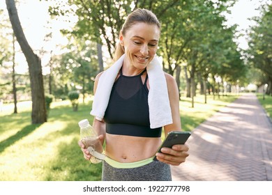 Energize your morning. Cheerful fit woman in sportswear refreshing, drinking water and using her smartphone after jogging in a green park on a sunny day - Powered by Shutterstock