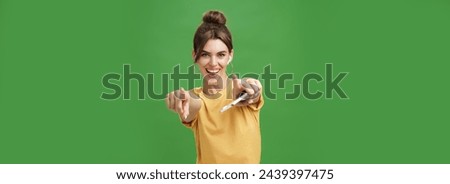 Energized charismatic attractive young woman with combed hair and cute diastema holding smartphone dancing joyfully listening music in wireless earphones pointing and smiling at camera over green wall
