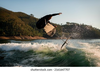 Energy Athletic Man In Black Wetsuit Jump With Foil Wakeboard Over Splashing Wave. Water Sports Activity.