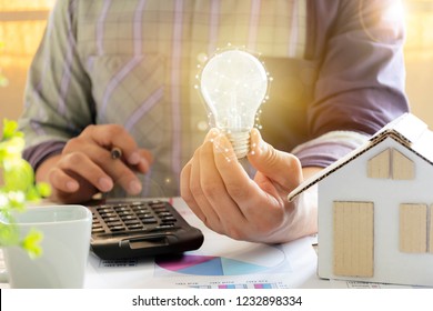 Energy Concept. Men Who Are Calculating Cost Savings From Energy. Hand Holding A Light Bulb, And A Pencil. Home Model, Calculator, Chart Document.